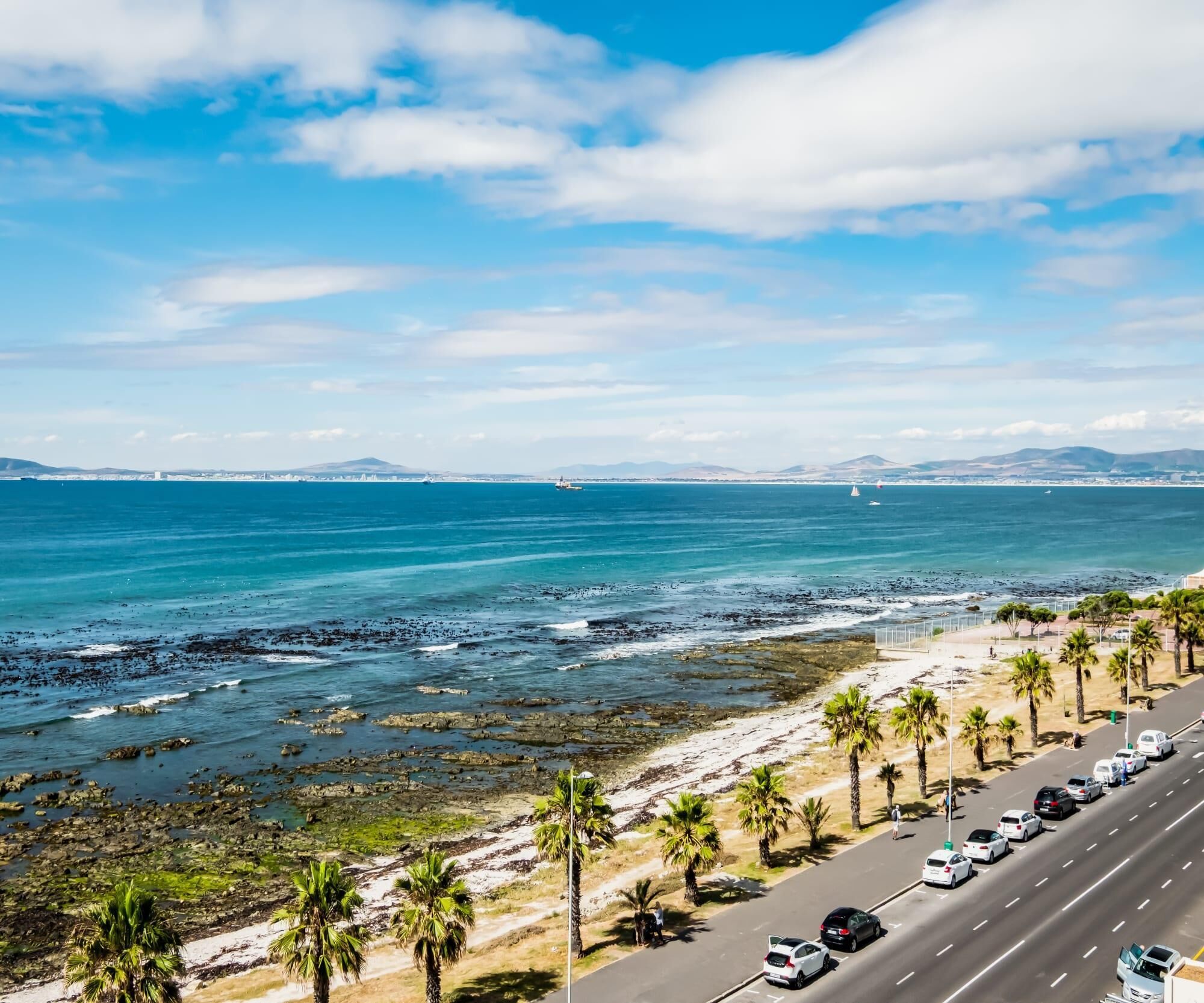 Una strada costiera con auto parcheggiate, palme e vista sul mare e montagne lontane.