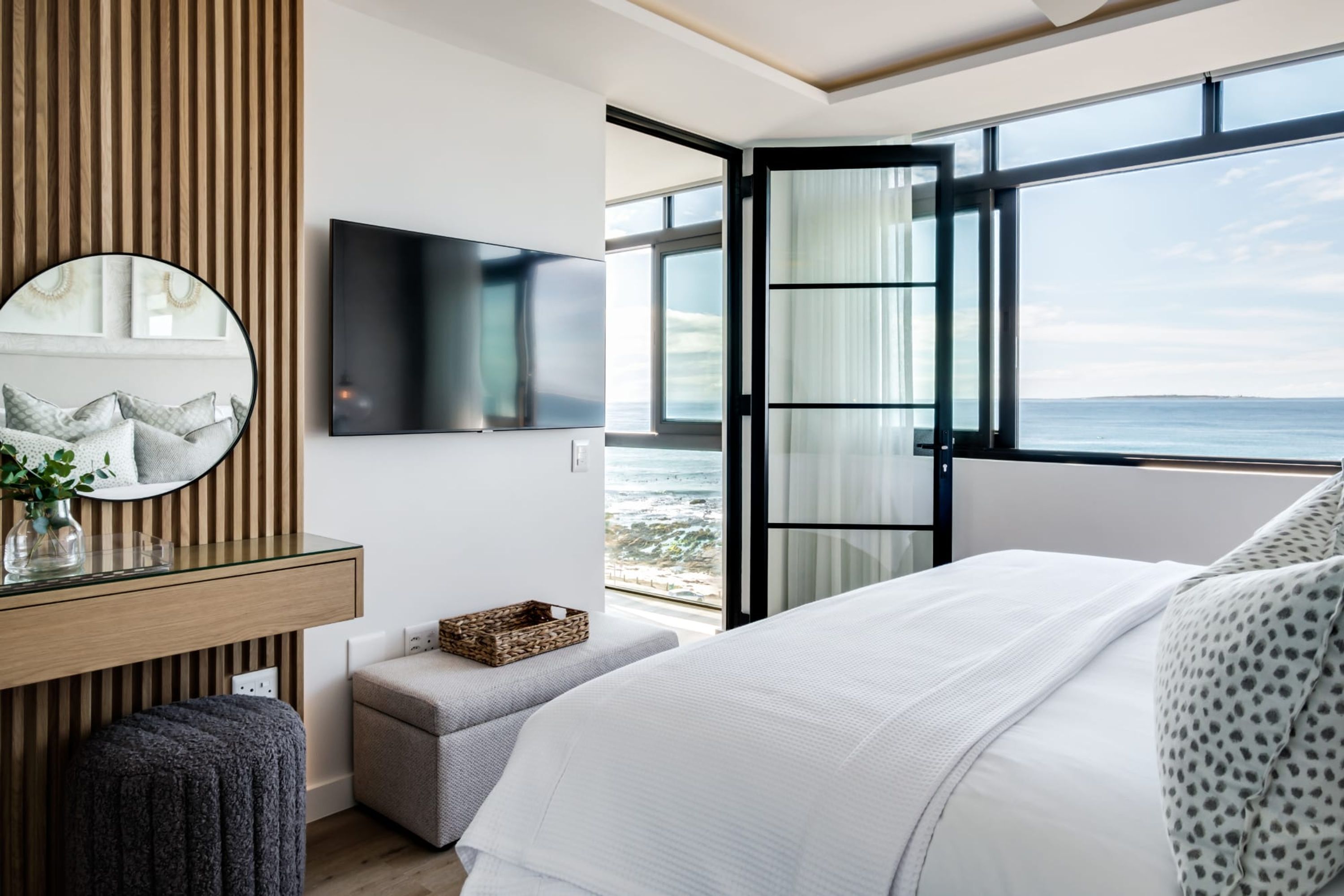 A modern bedroom with a large bed, a TV mounted on the wall, a wooden panel wall, and a view of the ocean.