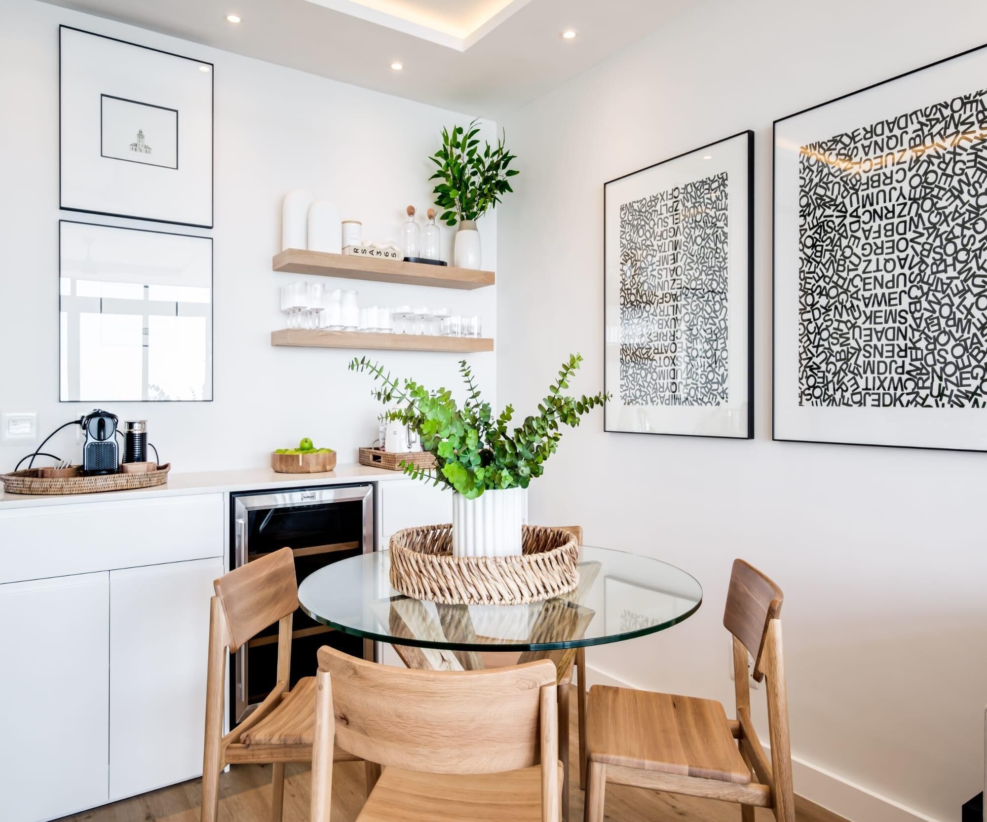 Eine moderne Küche mit weißen Schränken, einem Glastisch und Holzsesseln. An den Wänden befinden sich gerahmte Kunstwerke und auf der Arbeitsplatte ein Blumentopf.
