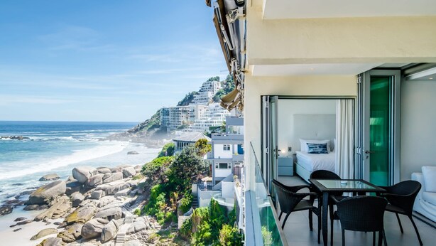 Ein Balkon mit Glastisch und Stühlen mit Blick auf einen Strand, der felsiges Gelände und Gebäude im Hintergrund zeigt.