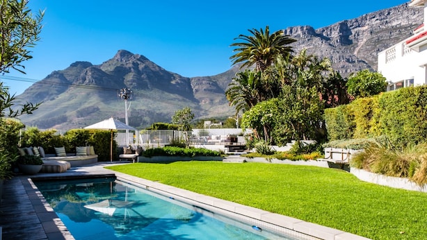 A swimming pool with a wooden deck, surrounded by a well-maintained lawn and a variety of plants. - Magnificent Heritage Home w Sweeping Views of the City Oranjezicht Heritage (Cape Town)