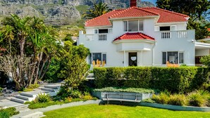 A two-story house with a red-tiled roof, surrounded by lush greenery and a mountain backdrop. - Magnificent Heritage Home w Sweeping Views of the City Oranjezicht Heritage (Cape Town)