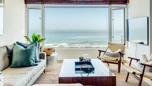 A modern living room with a large window overlooking the ocean, a comfortable sofa, a coffee table, and a TV. - Uninterrupted Ocean Views in Mouille Point Atlantic Vistas (Cape Town)
