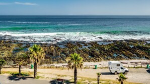 A beach with rocky shoreline, palm trees, a white RV, and a bench. - Uninterrupted Ocean Views in Mouille Point Atlantic Vistas (Cape Town)