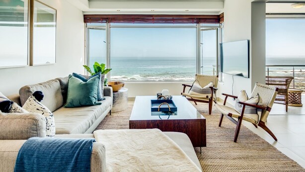 A modern living room with a large window overlooking the ocean, a comfortable sofa, a coffee table, and wicker chairs. - Uninterrupted Ocean Views in Mouille Point Atlantic Vistas (Cape Town)