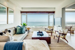 A modern living room with a large window overlooking the ocean, a comfortable sofa, a coffee table, and wicker chairs. - Uninterrupted Ocean Views in Mouille Point Atlantic Vistas (Cape Town)