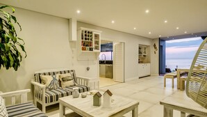 A spacious living area with a sofa, a coffee table, and a wicker chair. There is a view of the sea through an open door. - Perfect Family Holiday Villa in Camps Bay Spindrift (Cape Town)
