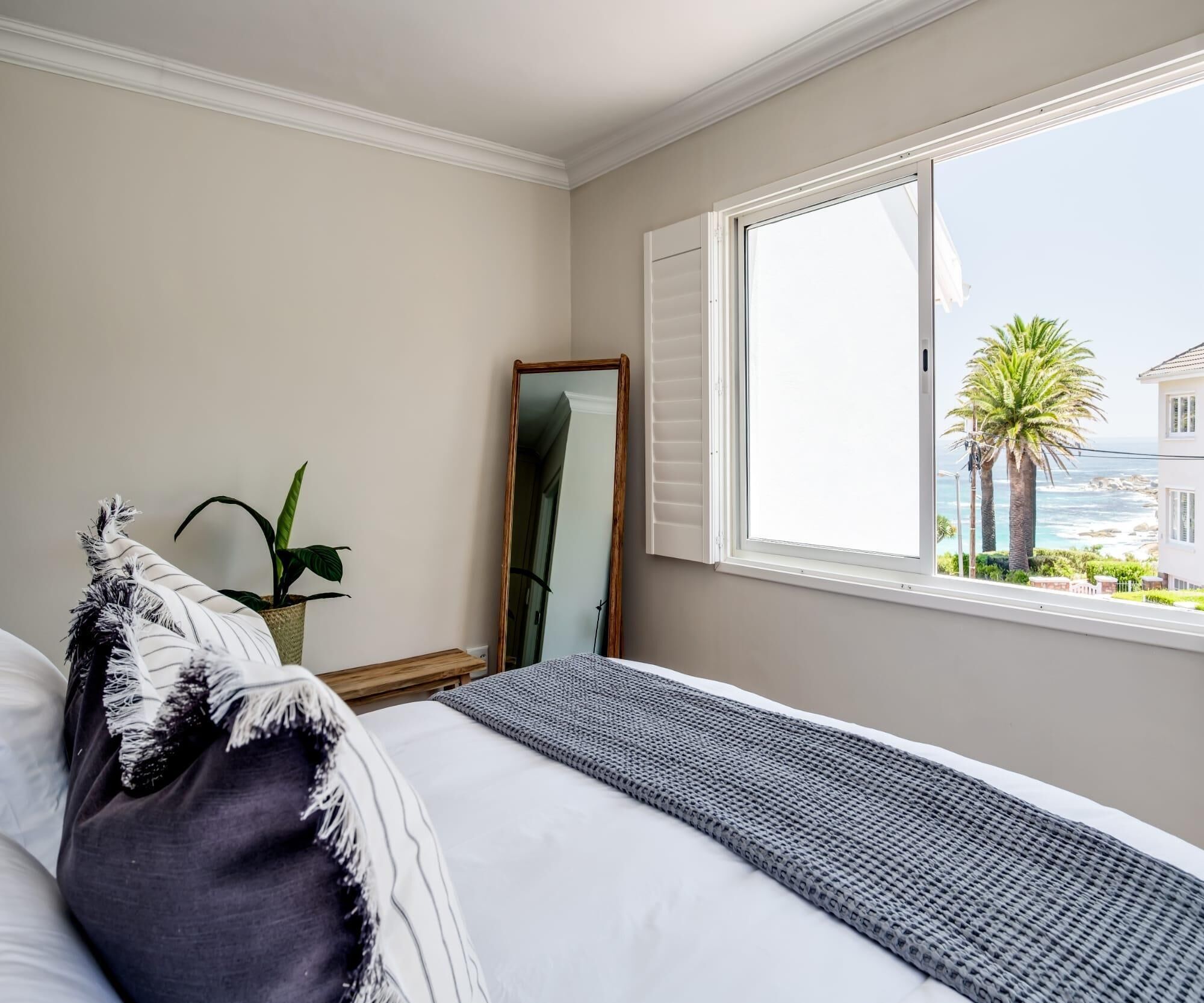 A bedroom with a bed, a window with a view of the sea, a potted plant, and a mirror.