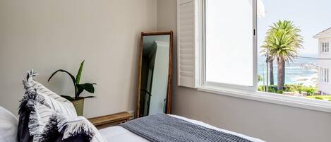 A bedroom with a bed, a window with a view of the sea, a potted plant, and a mirror.