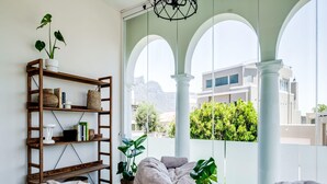 A modern living room with a balcony view, featuring a wooden bookshelf, two bean bags, and a round wooden table with a book. - Magical Holiday Home Walking Distance to the Beach Sedgemoor Villa (Cape Town)