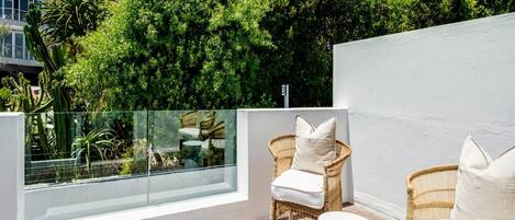 A wooden deck with wicker furniture and a glass balustrade overlooking a lush garden and mountains.