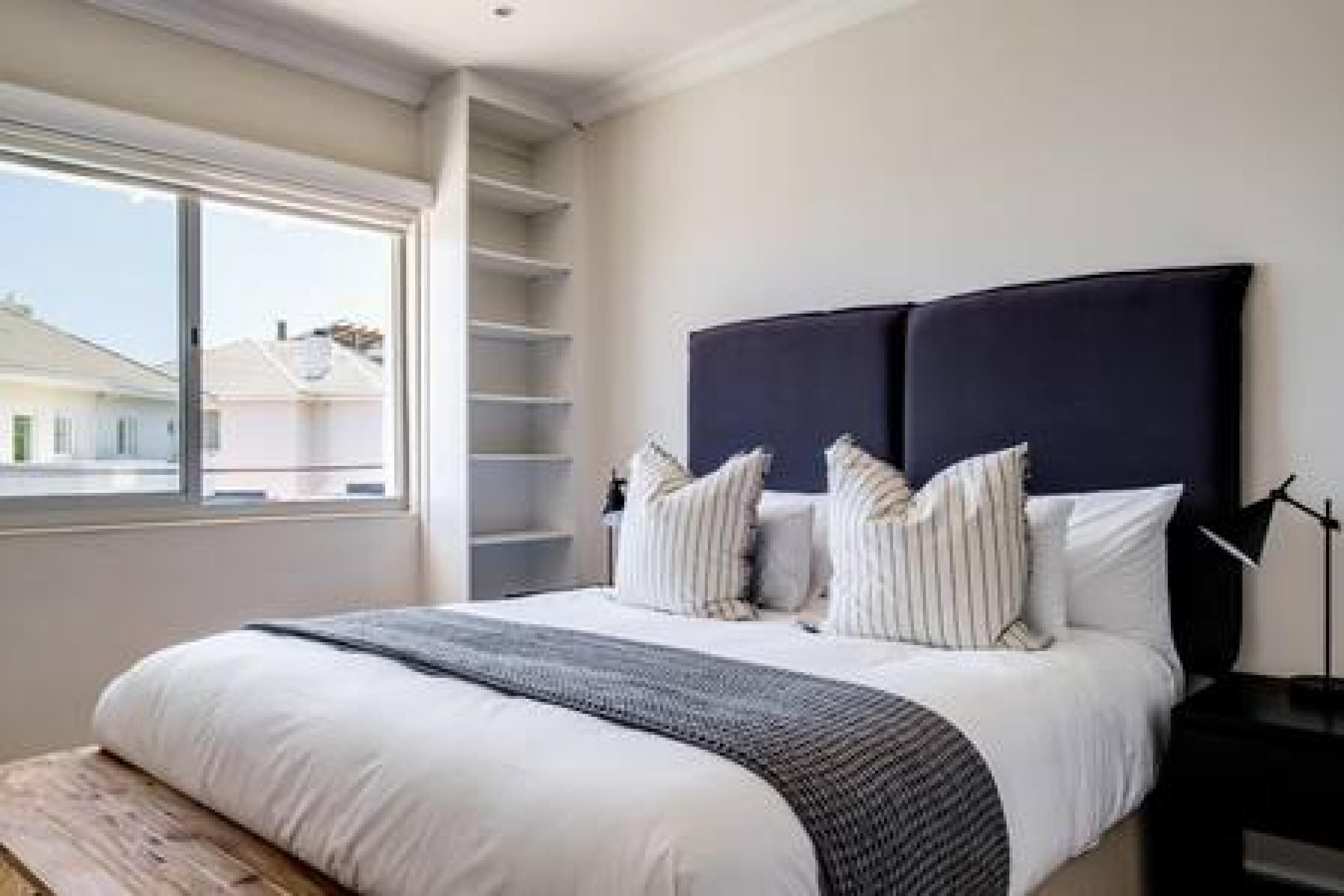 A bedroom with a bed, a bedside table, a lamp, and a window with a view of houses.
