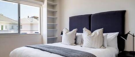 A bedroom with a bed, a bedside table, a lamp, and a window with a view of houses.