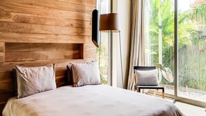 Ein modernes Schlafzimmer mit einem hölzernen Kopfteil, einem großen Bett mit beigefarbener Bettwäsche, einer Stehlampe und Blick auf Grün durch ein großes Fenster.