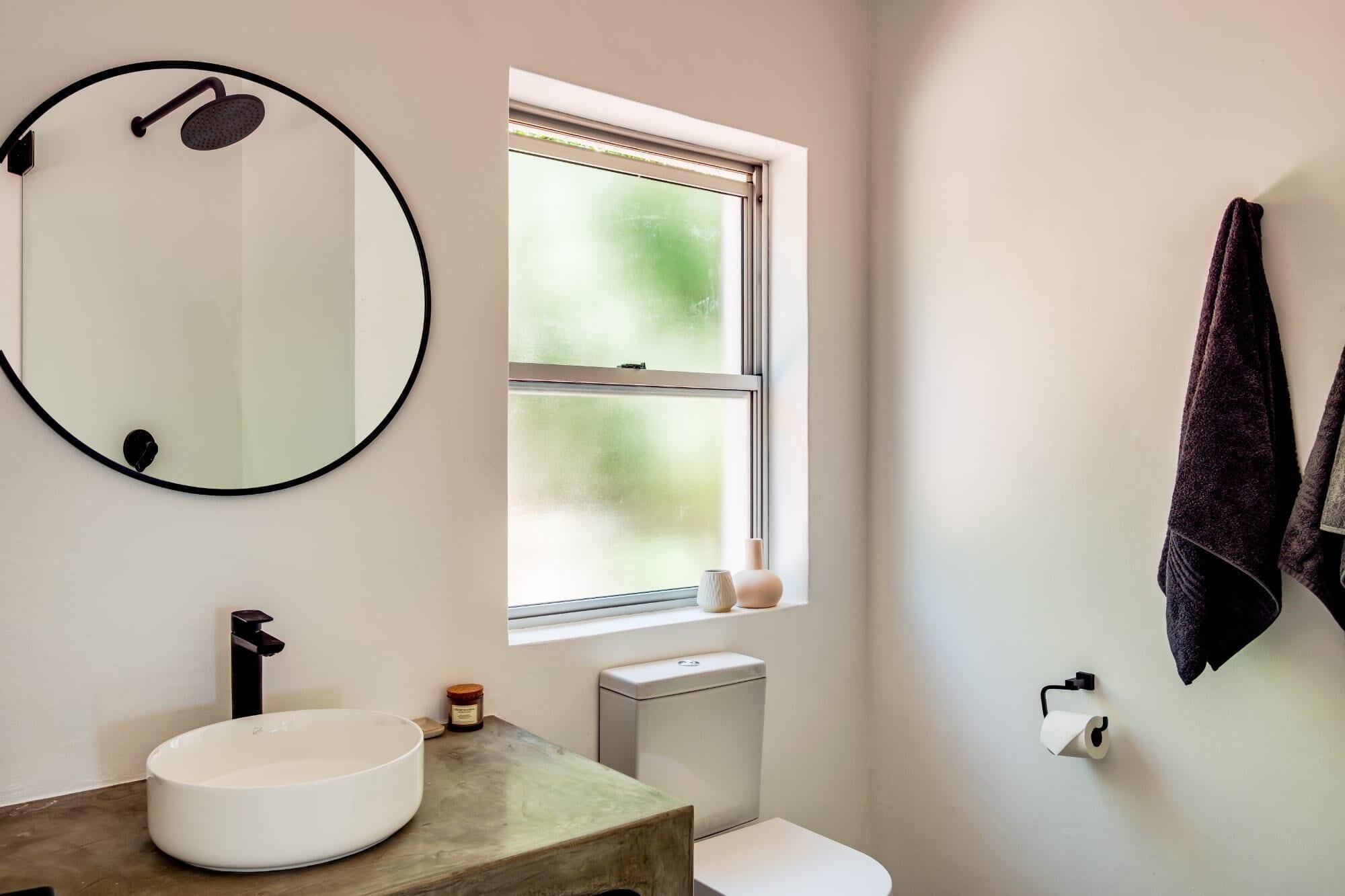 A bathroom with a round mirror, a sink, a toilet, and a window with a view of greenery.