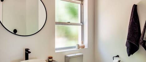A bathroom with a round mirror, a sink, a toilet, and a window with a view of greenery.