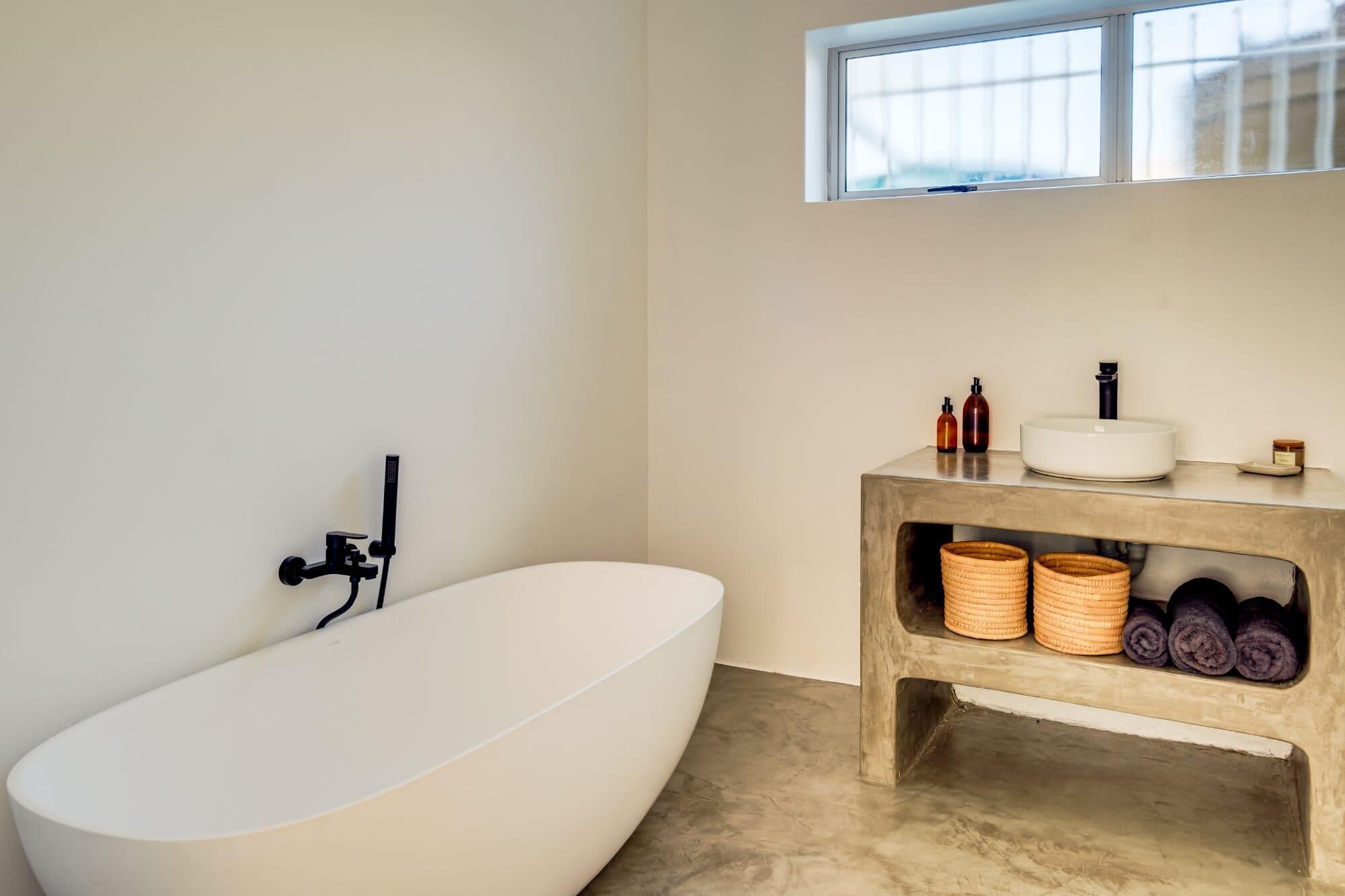 Een moderne badkamer met een vrijstaand bad, een houten wastafelmeubel met gevlochten manden, en een raam boven de wastafel.