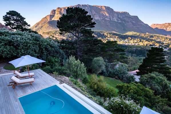 A swimming pool with lounge chairs and an umbrella, surrounded by lush greenery and a mountain view.
