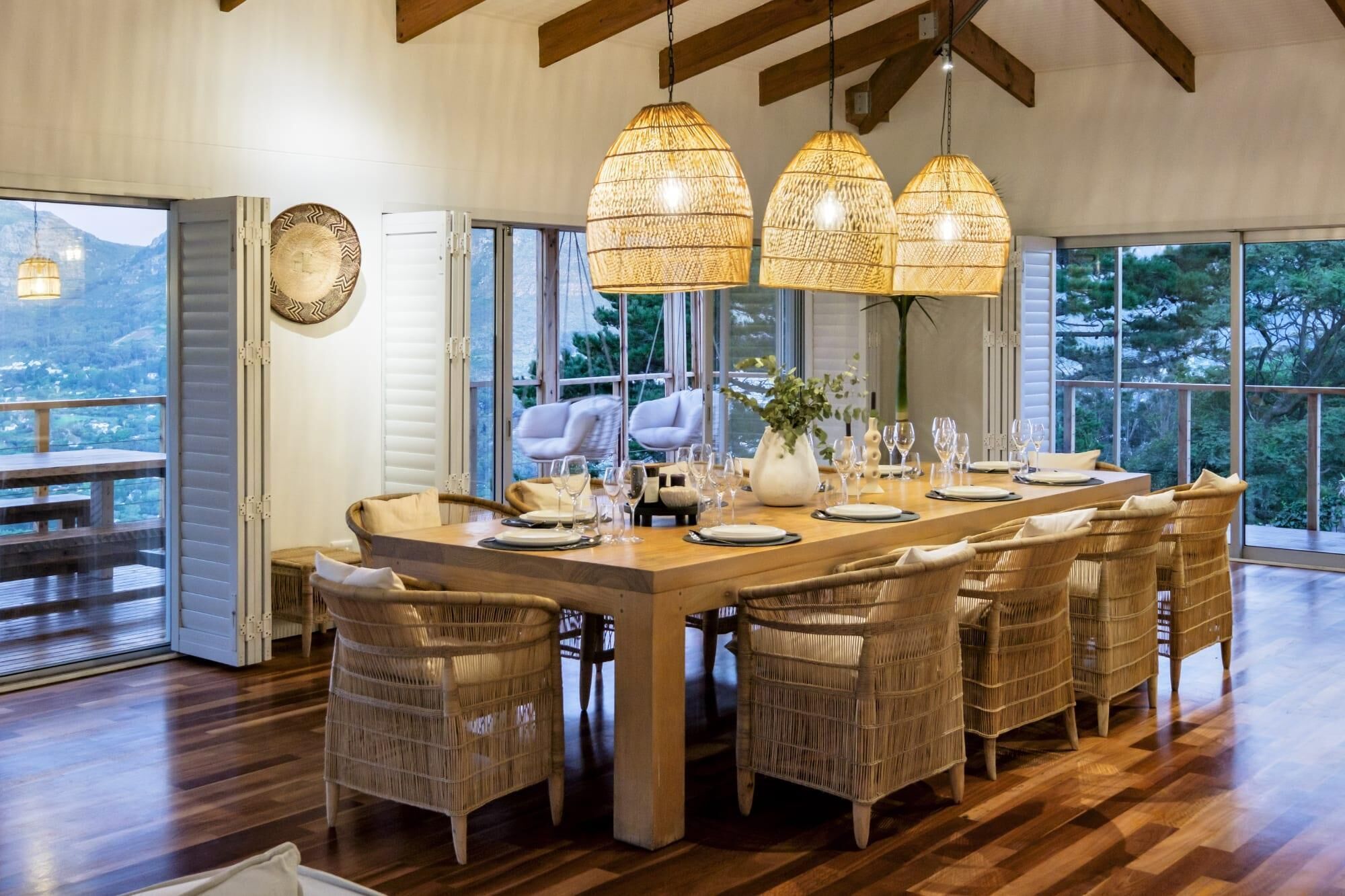 A dining area with a large wooden table set for six, surrounded by wicker chairs, under three hanging wicker lamps.