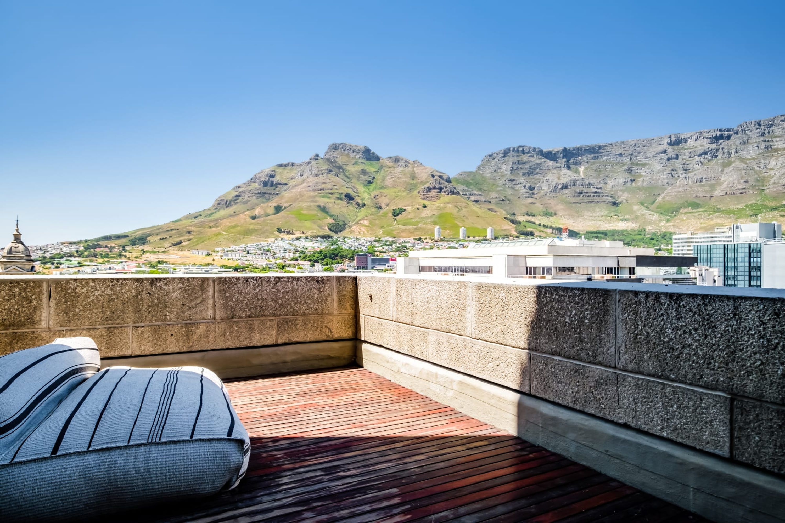 A rooftop terrace with a view of mountains, a stone wall, and a cushion.