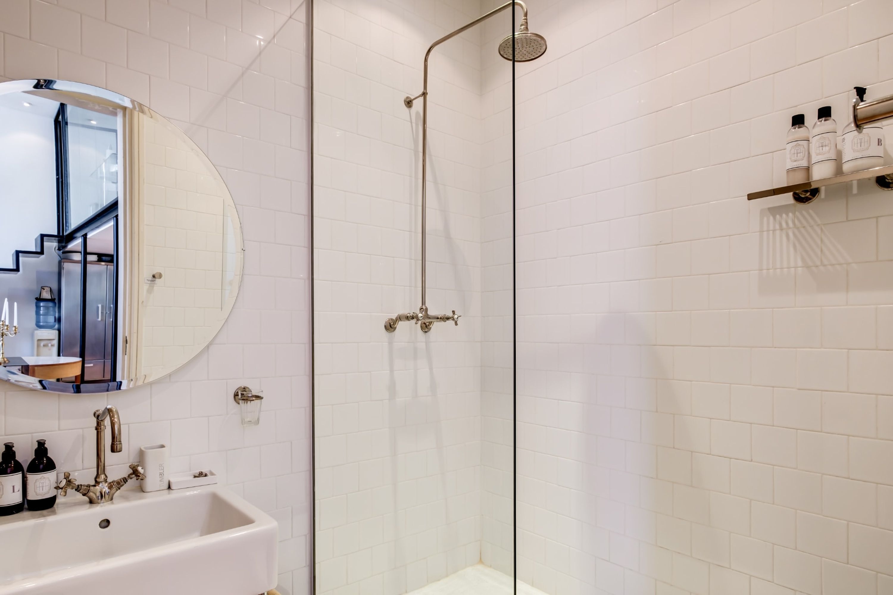 Ein Badezimmer mit weiß gefliesten Wänden, einem runden Spiegel, einem Waschbecken mit Wasserhahn und einer Duschkabine aus Glas.