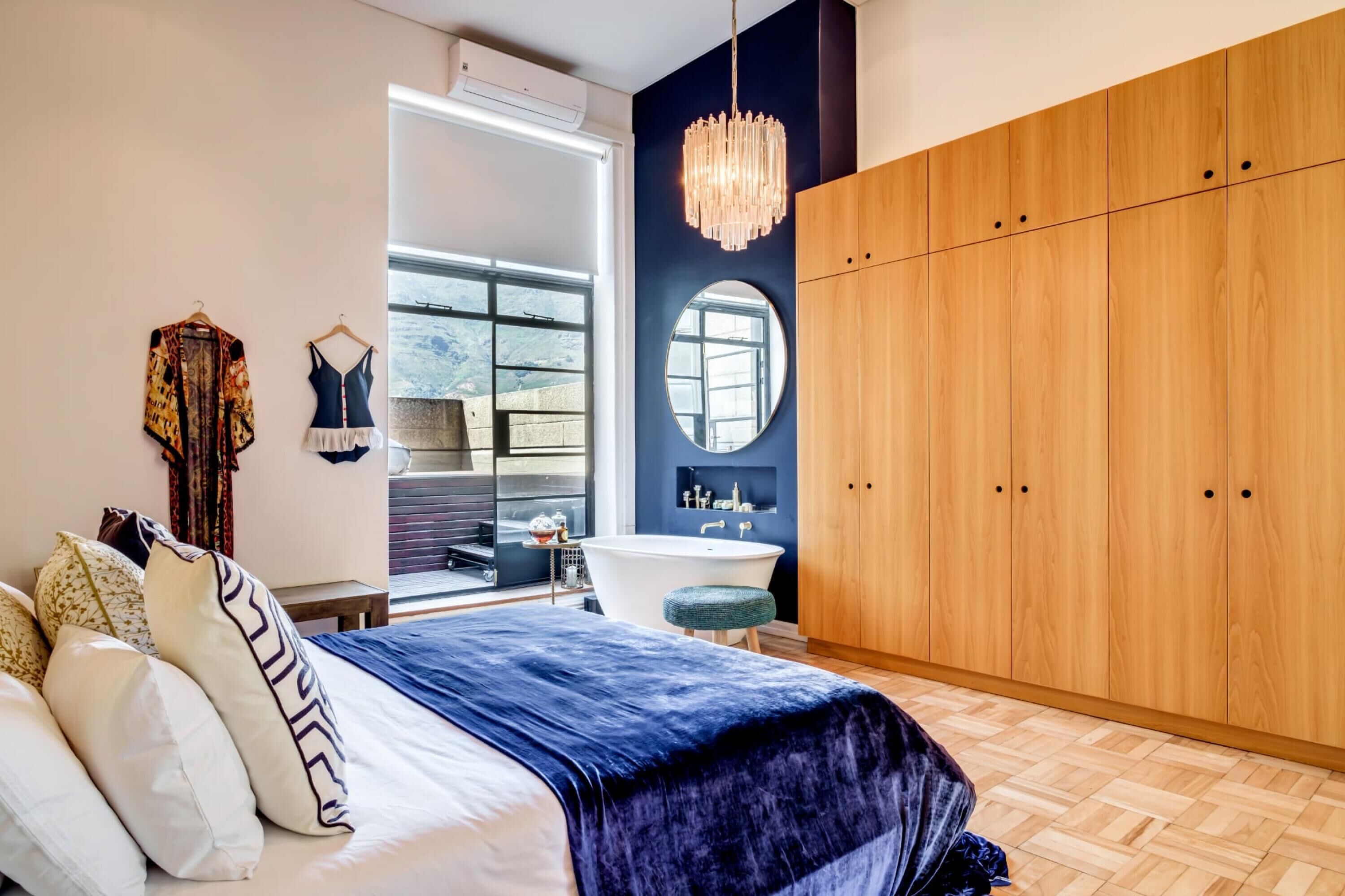 A bedroom with a bed, a large wooden wardrobe, a round mirror, a chandelier, and a view of the outdoors.