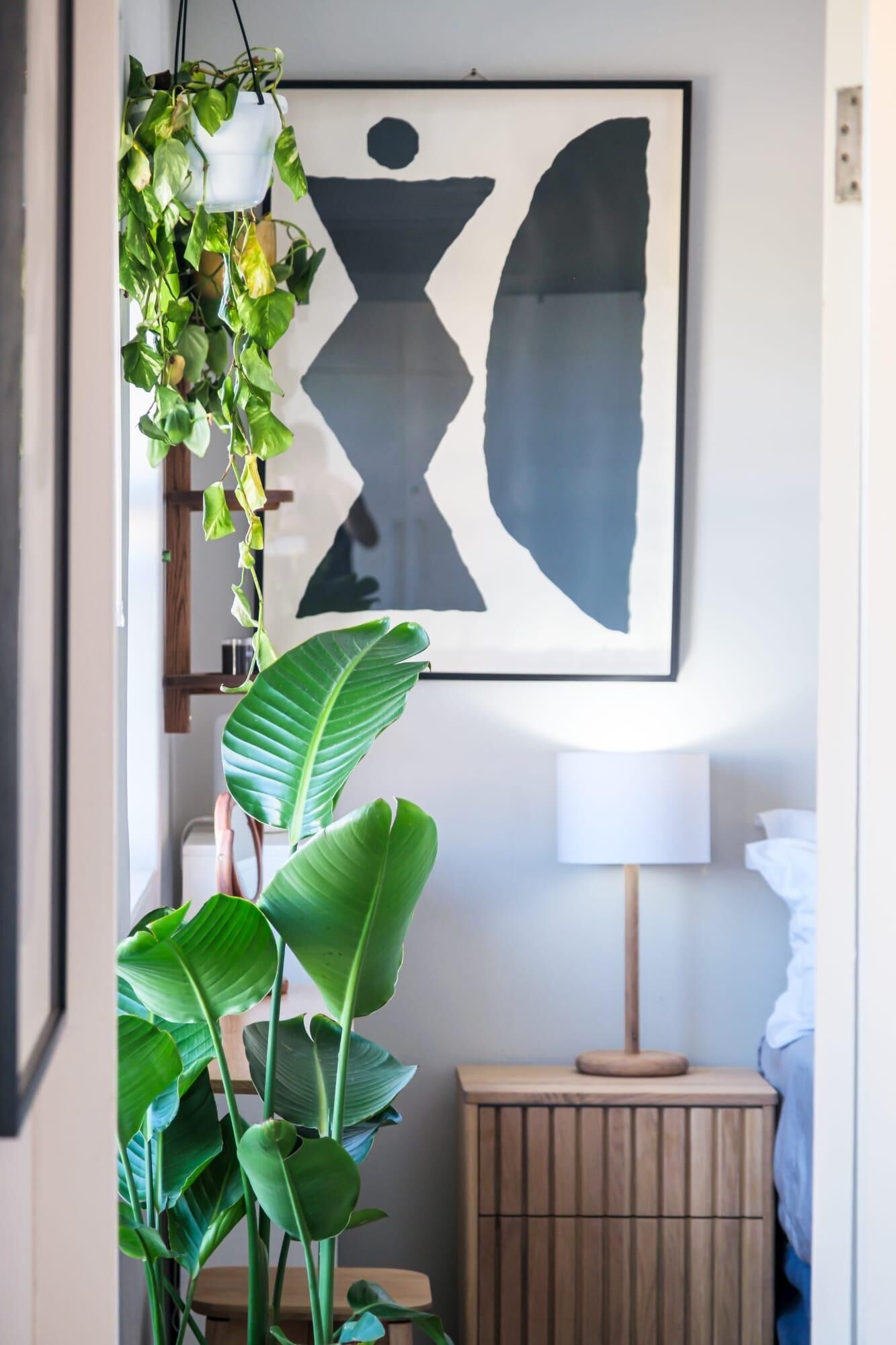A room with a large abstract painting, a potted plant, a bedside table with a lamp, and a wooden nightstand.