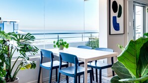 Une chambre moderne avec une grande fenêtre donnant sur la mer, une table blanche avec des chaises bleues et une plante en pot.