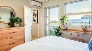 Une chambre moderne avec un grand lit, une commode en bois, un miroir et un balcon offrant une vue sur les bâtiments et la verdure.