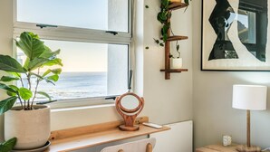 Une pièce avec un bureau placé près d’une fenêtre donnant sur la mer, une chaise en bois, une table de chevet avec une lampe et une étagère murale ornée de plantes.