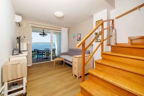 A living room with a wooden staircase, a sofa, a dining table, and a view of the sea through a glass door.