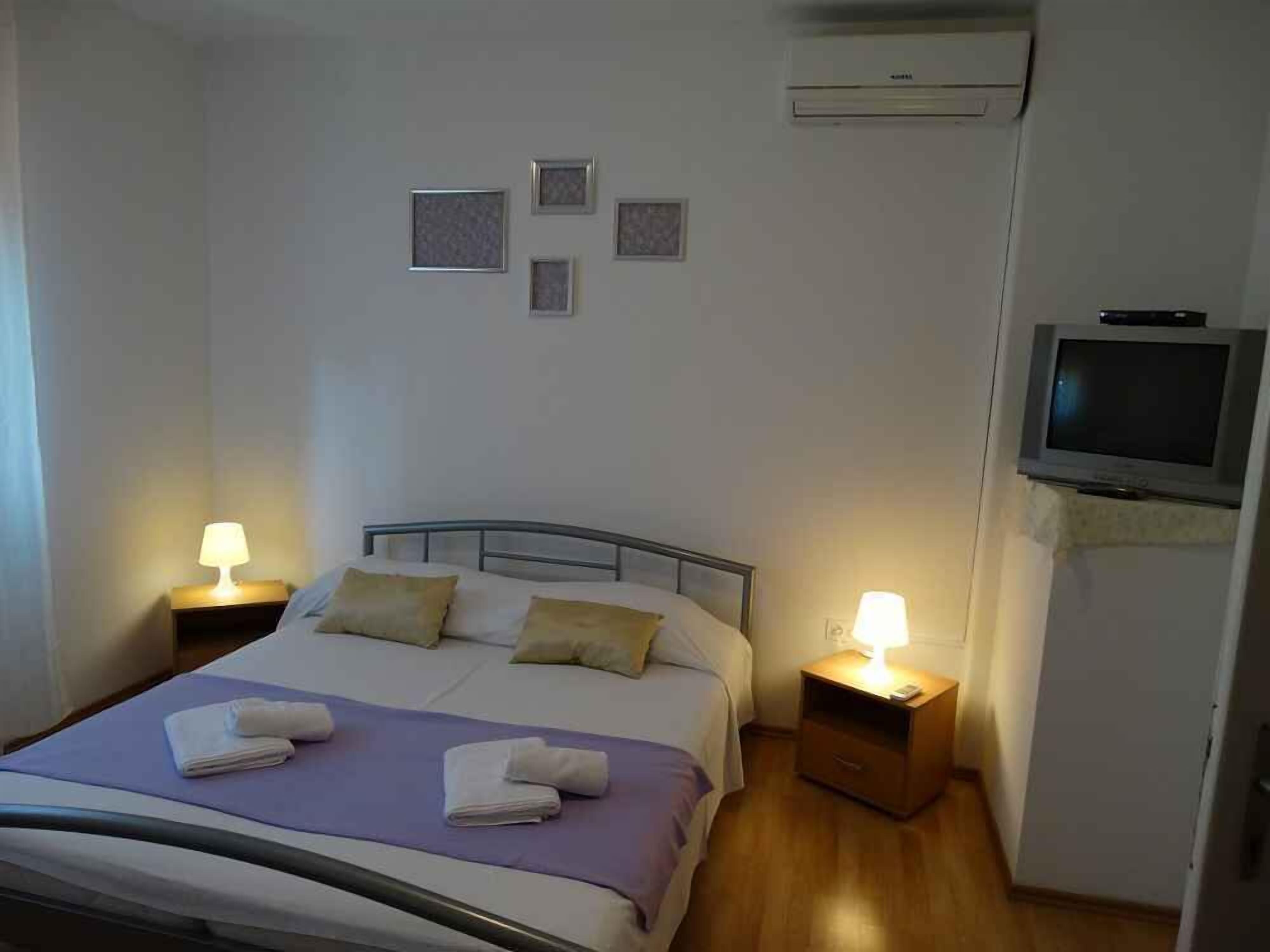 A neatly made bed with pillows and towels, two bedside tables with lamps, a television mounted on the wall, and a small air conditioning unit.
