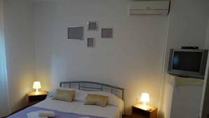 A neatly made bed with pillows and towels, two bedside tables with lamps, a television mounted on the wall, and a small air conditioning unit. - Holiday apartment with air conditioning (Crikvenica)