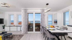 A modern dining area with a large table, chairs, and a view of the sea through glass doors.