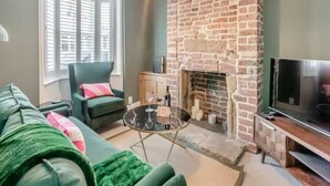 Un salon avec une cheminée en briques, un canapé vert, un téléviseur et une fenêtre avec des stores.