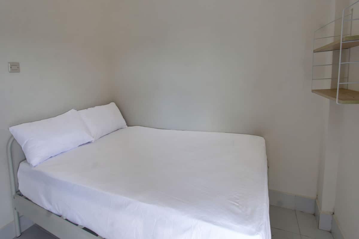Een kleine kamer met een enkel bed, een witte matras en kussens, een metalen bedframe, een witte muur en een klein houten plankensysteem.