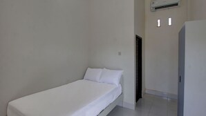 A small, single-bed room with a white bedspread, a white wall, and a white tiled floor. There is a white air conditioning unit mounted on the wall.