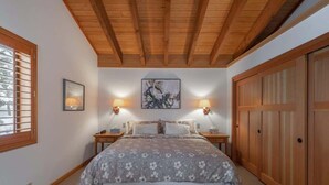 Ein Schlafzimmer mit Holzbalkendecke, einem Bett mit floralem Überwurf, zwei Wandlampen, einem Gemälde und einem Holzschrank.