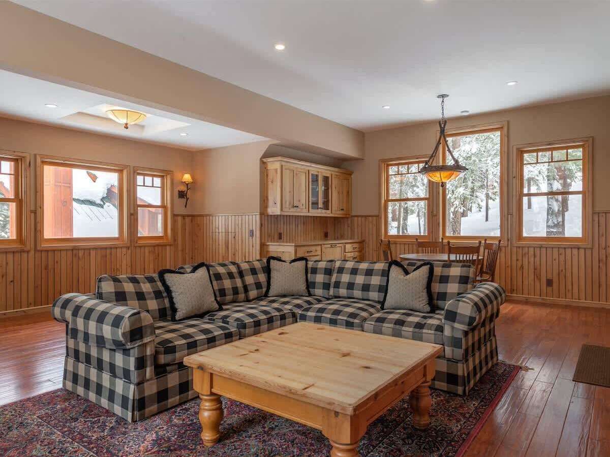 A large living room with a checkered sectional sofa, a wooden coffee table, and a kitchen area with wooden cabinets and large windows.