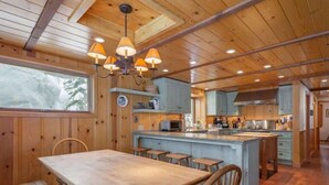 Ein Interieur einer Holzhütte mit einem Esstisch, Bänken, einer Küche mit Kochinsel und einer Deckenleuchte.