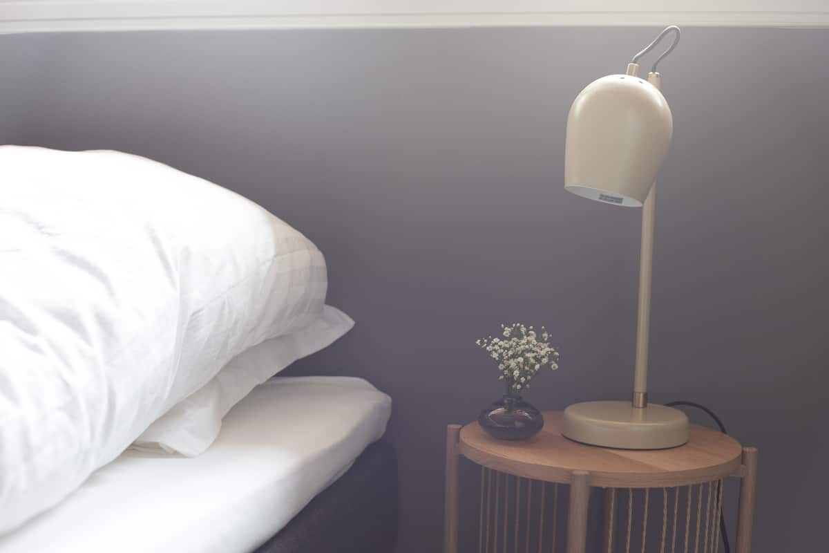 A bed with white bedding, a bedside table with a lamp, and a small vase with flowers.
