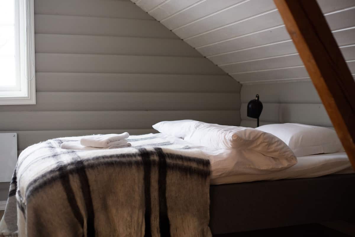 A bedroom with a sloped ceiling, a bed with white linens, a blanket, and a bedside table with a lamp.