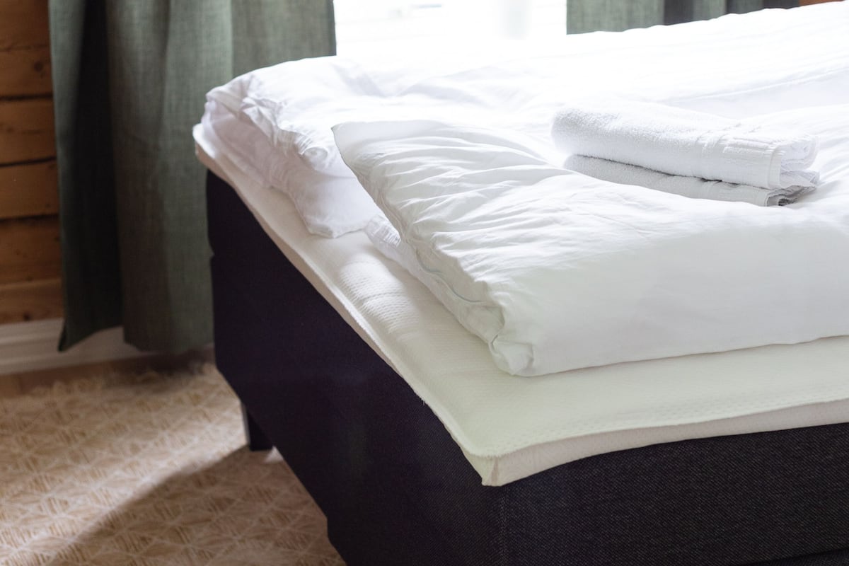 A neatly made bed with white linens and a folded towel, a dark-colored headboard, and a window with green curtains.