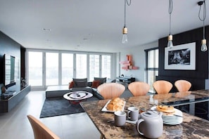 A modern living room with a large dining table, seating, and a view through large windows.