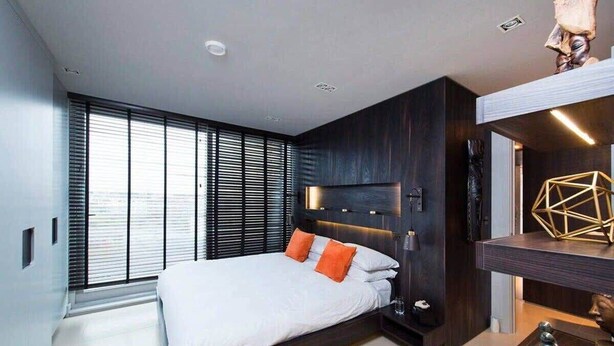Quarto moderno com cama, painéis de madeira escura, uma TV e uma escultura geométrica numa prateleira.