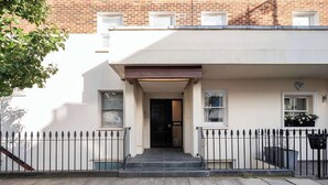 Una casa adosada con entrada principal, un pequeño porche y un edificio de ladrillo al fondo.