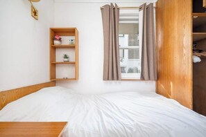 A single bed with a wooden headboard, a small wooden shelf with decorative items, and a window with curtains.