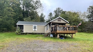 Une cabane à deux étages avec une terrasse, entourée d’une cour verdoyante et d’arbres.