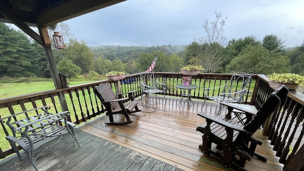 Une terrasse en bois, agréée de meubles d’extérieur, dont des fauteuils à bascule et une table, qui offre une vue sur un paysage verdoyant.