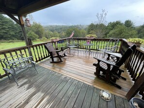 Une terrasse en bois, agréée de meubles d’extérieur, dont des fauteuils à bascule et une table, qui offre une vue sur un paysage verdoyant.